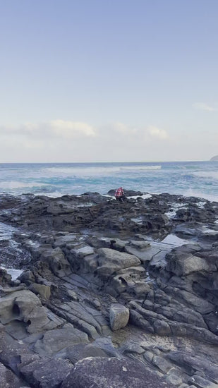 Scenic view of man taking photo on a rugged, rocky shoreline with gentle ocean waves breaking against the stones under a serene blue sky with distant clouds