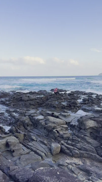 Scenic view of man taking photo on a rugged, rocky shoreline with gentle ocean waves breaking against the stones under a serene blue sky with distant clouds