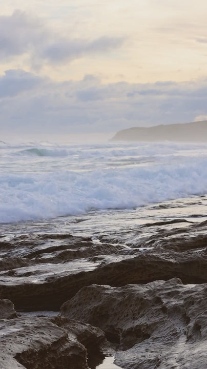 White ocean waves crashing on rocky coastline with glistening reflections at golden hazy sunset in slow motion