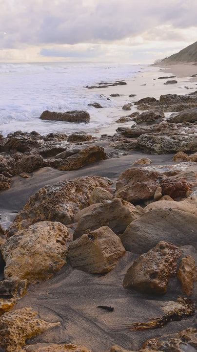 Soft, rhythmic ocean waves gently caressing dark volcanic sand and weathered rocks along an isolated coastline during early morning light in slow motion
