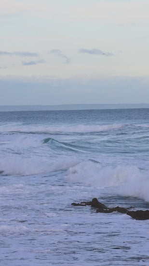 Metallic monochromatic blue ocean with soft waves crashing on rocky shoreline at sunset , creating serene seascape