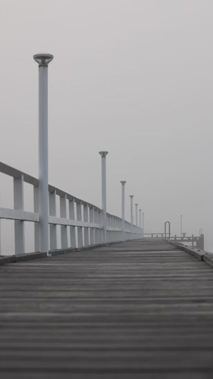Empty wooden pier with white railings and modern lamps vanishing into the distance on a moody, foggy morning. Gloomy and melancholic scenery