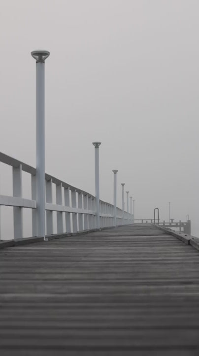 Empty wooden pier with white railings and modern lamps vanishing into the distance on a moody, foggy morning. Gloomy and melancholic scenery
