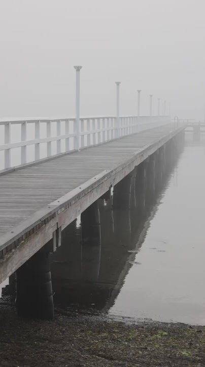 Mysterious long wooden pier extending into the calm water of a lake or sea, vanishing into the dense morning mist on an overcast day