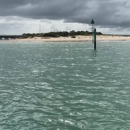 Light rain creating ripples on the surface of an inlet on a cloudy, overcast day. A distant bridge and a sandy shore visible in the background