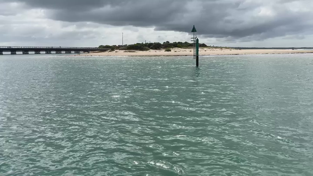 Light rain creating ripples on the surface of an inlet on a cloudy, overcast day. A distant bridge and a sandy shore visible in the background