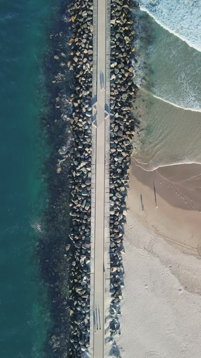 Stunning top down aerial shot captures a stone jetty at at The Spit, Gold Coast separating the calm turquoise sea from a sandy beach with rolling waves and people walking