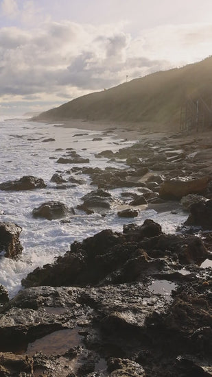 Gentle waves washing over dark rocks on the coast at dawn, with a green hill in the background in slow motion
