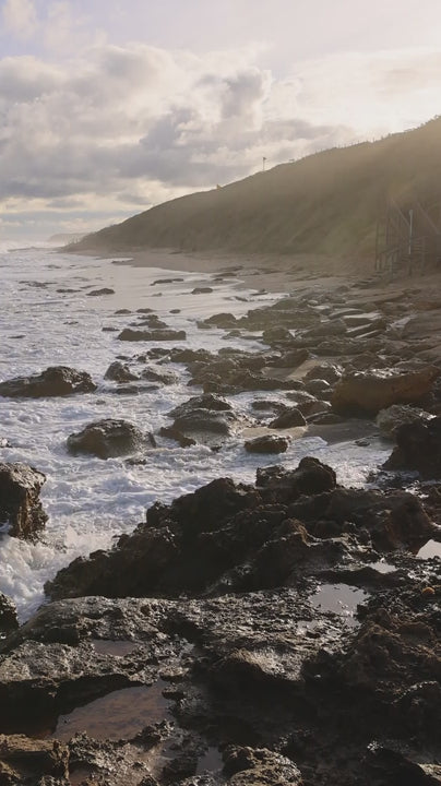 Gentle waves washing over dark rocks on the coast at dawn, with a green hill in the background in slow motion