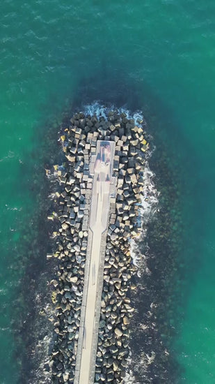 Aerial top down view of a stone breakwater extending into the turquoise ocean. People are walking on the pier as gentle waves lap the shore at The Spit, Gold Coast