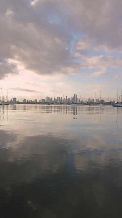 Melbourne urban skyline reflecting on glassy water during golden sunset, revealing sailboats against cloudy sky with serene coastal atmosphere