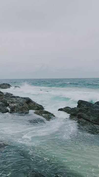 Dramatic ocean waves crashing against rugged black rocks on a cloudy, moody day. Beautiful and powerful turquoise water swirling around the shore