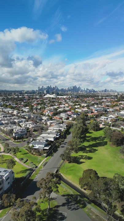 Aerial of Melbourne's suburbs stretch out beneath a blue sky, revealing a mix of traditional and modern homes, parks, and the city's skyline in the distance