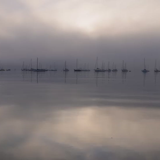 Serene and moody scene of sailboats moored in a calm harbor on a foggy morning. The sun breaks through the thick mist, reflecting on the still water