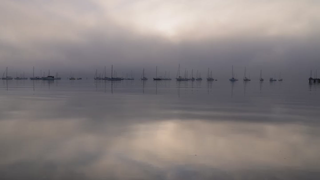 Serene and moody scene of sailboats moored in a calm harbor on a foggy morning. The sun breaks through the thick mist, reflecting on the still water