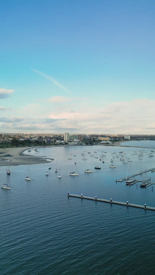 Fly over marina with boats docked in St. Kilda harbor towards St Kilda with blue sky and calm water