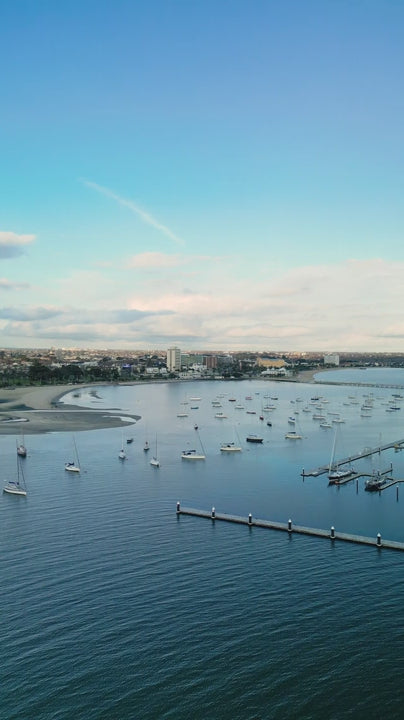 Fly over marina with boats docked in St. Kilda harbor towards St Kilda with blue sky and calm water