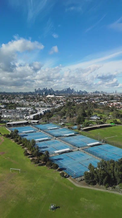 Aerial of Melbourne blue outdoor tennis courts with city skyline view in the distance with blue sky, green trees, and surrounding residential landscape - fly over