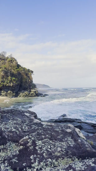 Beautiful view of ocean waves gently breaking on a rocky shore. A rugged cliff covered in green vegetation overlooks the tranquil sea