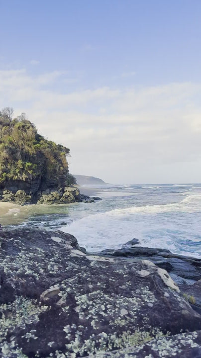 Beautiful view of ocean waves gently breaking on a rocky shore. A rugged cliff covered in green vegetation overlooks the tranquil sea