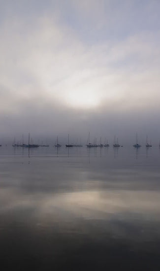 Hazy morning view of a misty ocean with sailboats in the distance. Still water perfectly reflecting the foggy sunrise and moody sky