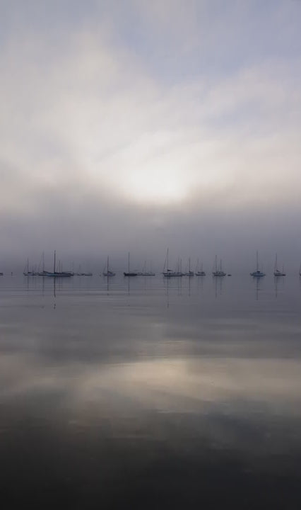 Hazy morning view of a misty ocean with sailboats in the distance. Still water perfectly reflecting the foggy sunrise and moody sky